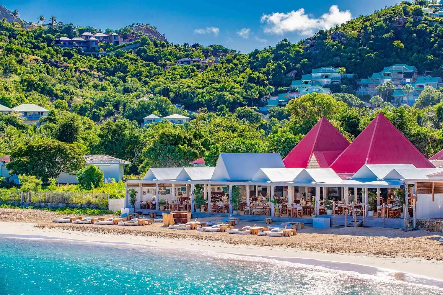 A beach restaurant with bright peaked roofs on the sand of St Jean, villas on the hill behind