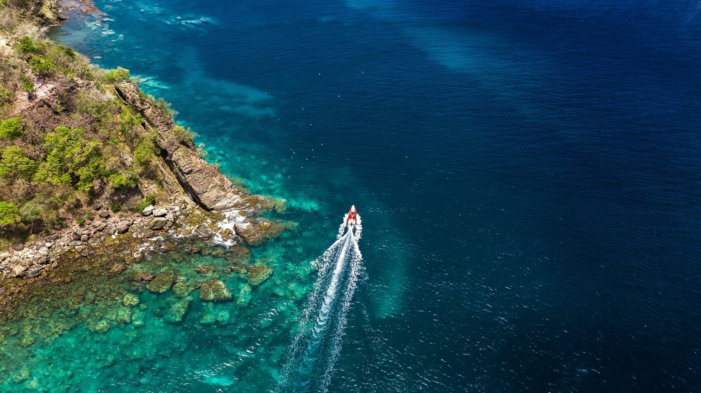 A boat leaving a white wake along the rocky coast, seen from above