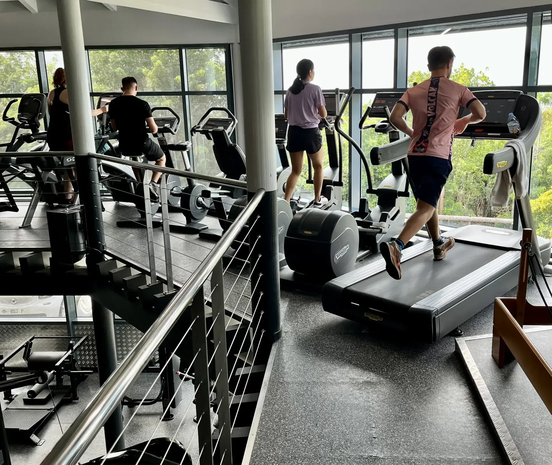 Des personnes à l’entraînement sur tapis de course et machines dans une salle de sport lumineuse et vitrée à Saint-Barth, la végétation derrière les baies