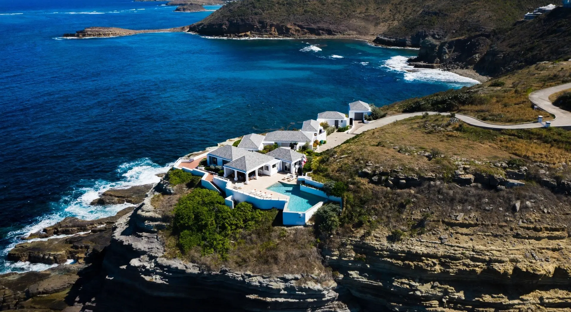 A white villa on a rocky point seen from the air, waves breaking below